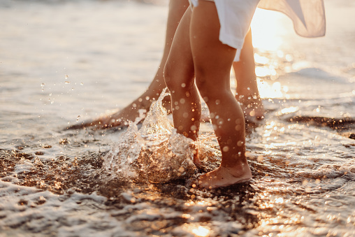 The bare feet of an adult woman and a girl, a child in white dresses are splashing in the ocean at sunset. Water drops sparkle in the sun
