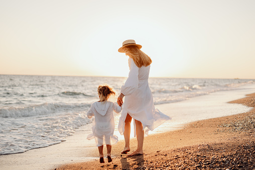 A young girl, mother, with a little daughter in white dresses, walk on the water on the seashore at sunset.