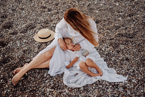 A young mother and daughter in white dresses are sitting on a rocky ocean shore. The little daughter lay down on her mother and looks into the camera.