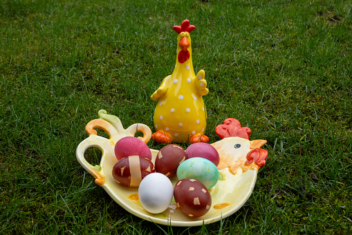 Ceramic rooster near dyed easter eggs on the chicken-shaped plate on green background. Easter celebration.