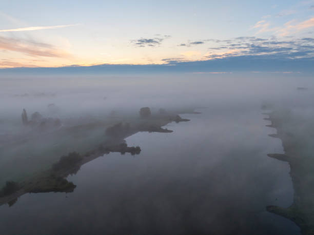 River IJssel covered in a blanket of mist seen from above during a fall morning near the river IJssel in Overijssel. The treetops are popping out over the thick fog with some of the trees starting to change color during the annual autumn season. The fog is giving the landscape a mystical atmosphere.