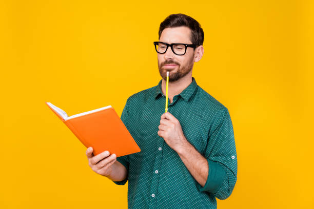 foto de homem inteligente intrigado vestido camisa pontilhada em óculos olhar cuidadosamente para lápis de livro no queixo isolado no fundo de cor amarela - mid adult men smart casual white happiness - fotografias e filmes do acervo