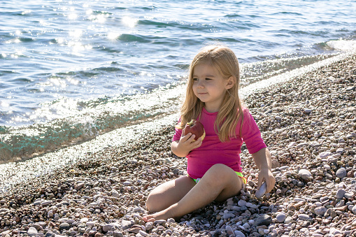 Young girl with long blond hair, dressed in a pink swimsuit eating a fresh apple on a sunny day at the beach with ocean view.
