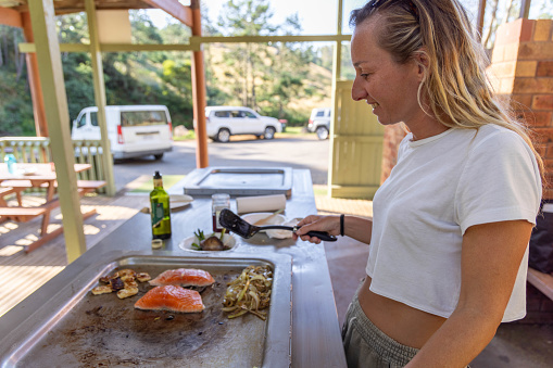 A vibrant young woman embraces the Australian outdoor lifestyle as she skillfully tends to a sizzling barbecue, infusing the air with the enticing aroma of grilled delights. Enjoying the warmth of the sun, she captures the essence of leisurely outdoor cooking in the heart of Australia.