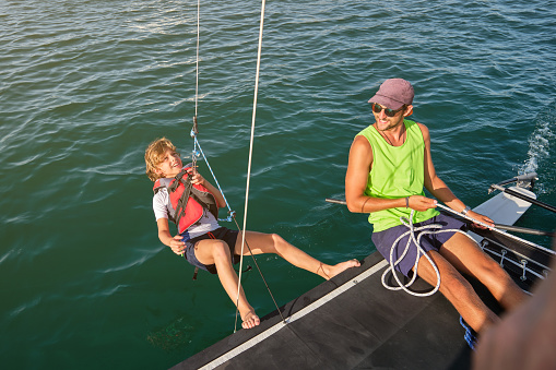 From above of cheerful young ethnic man in casual clothes and sunglasses smiling while pulling rope with fastened happy preteen boy during trip in sea on catamaran