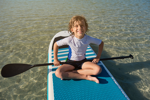 Full body portrait of smiling boy with paddle sitting on SUP board and looking at camera in middle of sea during summer vacation