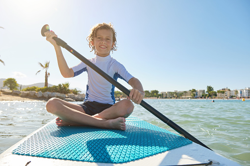 Full body of smiling preteen boy with wet hair sitting on paddleboard with crossed legs and rowing while floating in rippling sea against cloudless sky at resort