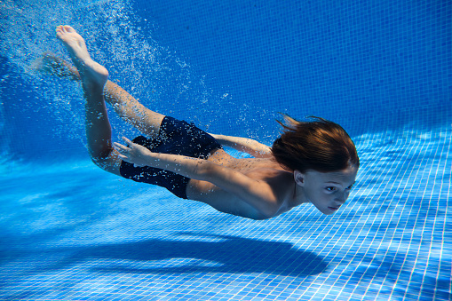 Side view of boy surrounded with air bubbles swimming near bottom of tiled pool on weekend summer day