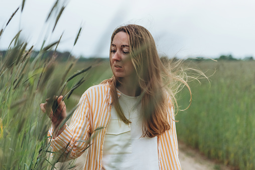 A girl, a woman stands in the middle of a field of green wheat. Touches ears of wheat with his hands. Summer cloudy day. The wind blows the girl's hair.