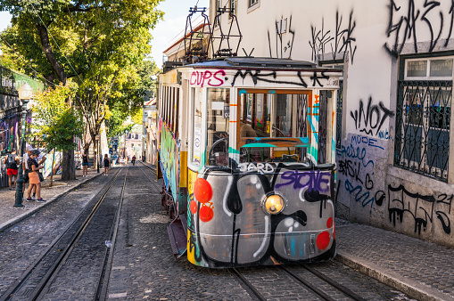 Lisbon, Portugal - January 5, 2023: A tramway surrounded by tall buildings in the daytime, with a yellow tram bus carrying passengers traveling along it. Two cars can also be seen moving in the opposite direction.