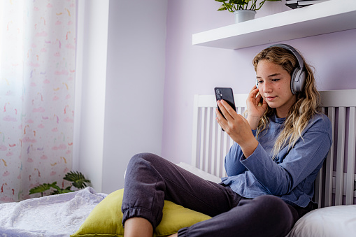 Teenager girl using headphones and text messaging on phone in her bedroom. High resolution 42Mp studio digital capture taken with SONY A7rII and Zeiss Batis 25mm F2.0 CF lens
