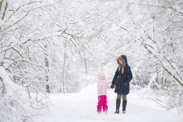 istock Smiling mother and daughter walking on white fresh snow covered road through tree branches at park after blizzard. Spending time together in beautiful winter day. Enjoying nature stroll. Front view. 2045238301