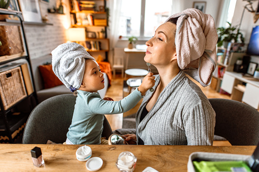Smiling young woman and little daughter with towels twisted around heads, looking at mirror during skincare procedure at home