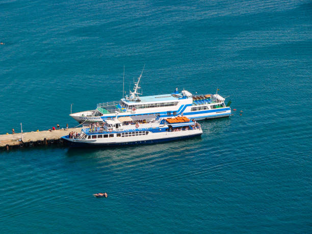 Two ships are moored near a small bay. All around is smooth blue sea water and a blue clear sunny sky.