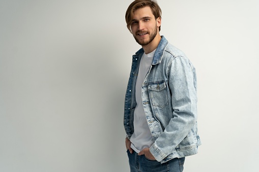 Close up studio portrait of stylish brutal young bearded European man wearing jeans jacket, posing at white blank wall with copy space for your text or promotional content