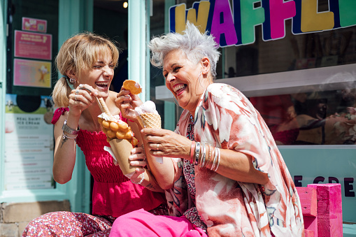 Two women sitting side by side on a bench outside of a cafe store eating ice cream and waffles together. They are enjoying the sunshine, laughing and talking together. They are exploring the city centre in Durham, North East England.