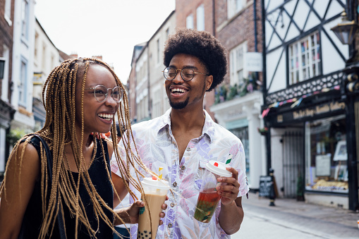 Two young adults enjoying time together on a city street drinking bubble tea together. They are enjoying the sunshine, laughing and talking together. They are exploring the city centre in Durham, North East England.