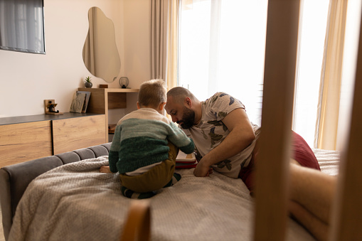 Father and son sitting on bed and talking to each other. Father and smiling little boy playing while sitting on bed. Portrait of happy child listening his father at home.