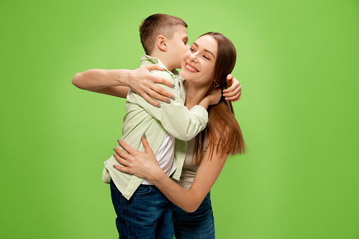 Happy, smiling little boy, son hugging and kissing his beautiful, caring mother, showing his love against green studio background. Concept of happiness, Mother's day, childhood, fashion and lifestyle