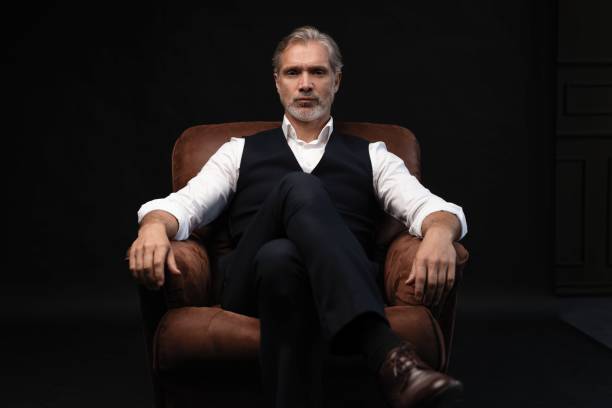 In leather chair sitting senior businessman with gray beard wearing dark suit. Against black wall In leather chair sitting senior businessman with gray beard wearing dark suit. Against black wall. old rich white guy stock pictures, royalty-free photos & images