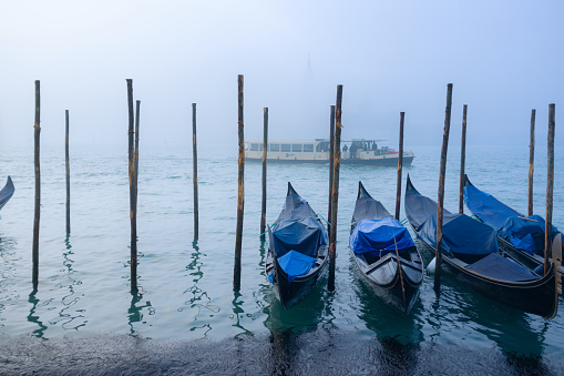 Gondolas tied up on the side of a misty Grand Canal in Venice, whilst beyond a vaporetto makes its way to the next destination. Across the water we would, on a clearer day, see the iconic shape of the Church of San Giorgio Maggiore