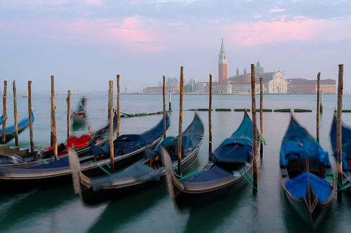 A beautiful sunset illuminates a line of gondolas tied up on the side of the Grand Canal in Venice. Across the lagoon we see the iconic shape of the Church of San Giorgio Maggiore.