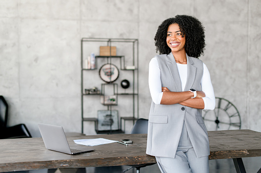 A joyful African-American businesswoman leaning on a desk in office space, her attire and demeanor exuding professionalism and modern elegance. The concept focuses on the harmony of business and style