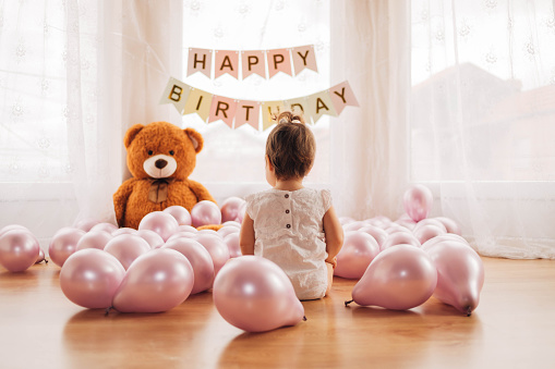 An adorable little girl is sitting on the floor and playing with purple balloons. The baby has lots of balloons and a big teddy bear on her first birthday.