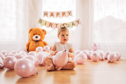 An adorable little girl is sitting on the floor and playing with a purple balloon. The baby has lots of balloons and a big teddy bear on her first birthday.