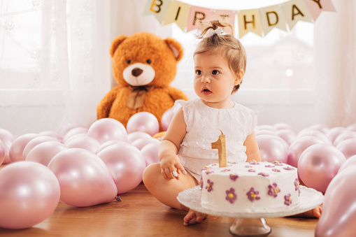 The little girl got the cake for the first time, so , she is sitting on the ground surrounded by balloons and looking at her birthday cake.