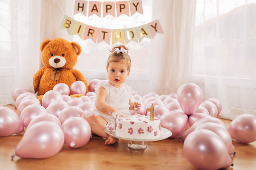 The adorable baby girl is sitting on the floor, surrounded by balloons and teddy bear, and touching the whipped cream from the birthday cake.