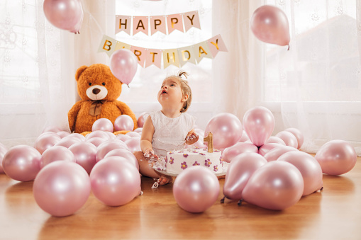 The cute baby girl is enjoying her birthday party. The child is sitting on the floor, eating the cake with her hands and looking at the balloons  falling from the ceiling