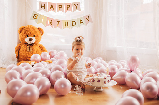 The adorable little girl is sitting on the floor and smashing her first birthday cake. The child is playing and eating the cake with her hands. Smash cake.
