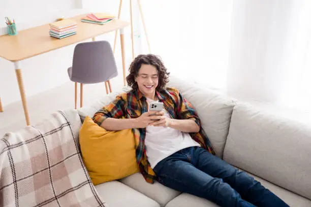 Photo of funky cheerful guy wear plaid shirt lying sofa texting instagram twitter telegram facebook indoors room home house.