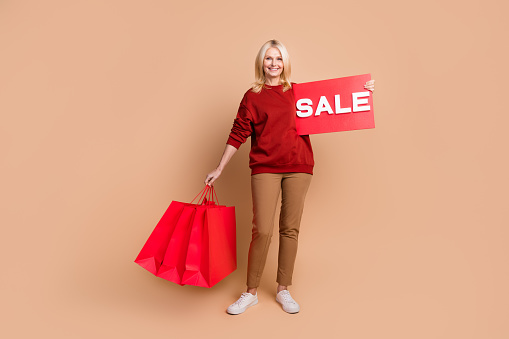 Full length photo of cheerful good mood woman wear red sweater holding sale billboard shoppers isolated beige color background.