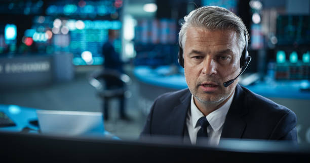 istock Middle Aged Stock Exchange Banker Working on a Desktop Computer with Real-Time Stocks, Commodities and Exchange Market Charts. Professional Investor Talking on a Mic with Business Partners 2045432897