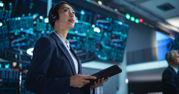 istock Young Asian Female Broker Working on Tablet Computer on Stock Exchange Floor. Traders Monitoring Real-Time Stocks, Commodities and Exchange Market Data. Professional Investment Agents in Office 2045455513