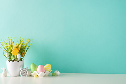 Spring Easter display: side view of a table with a floral shell pot, grass, tulips, a ceramic bunny, a container filled with rainbow eggs, and a clock, set against a gentle turquoise wall