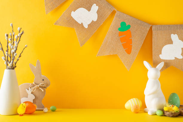 istock Easter theme setup, showcasing side view of tabletop adorned with rabbits, vibrant eggs, chicks, vase of pussy willows, a country-style garland against orange backdrop, leaving space for text or ads 2045456660