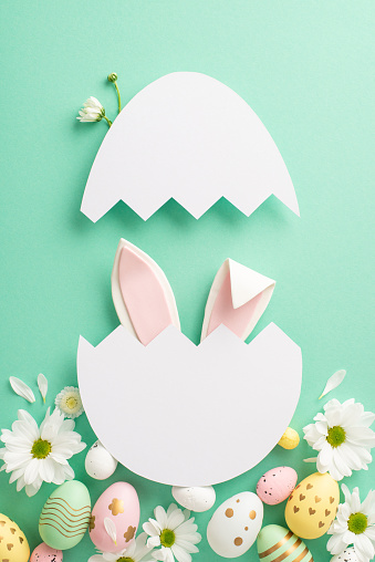 Easter charm layout. Top view vertical image of adorable hare ears visible from a broken eggshell, colored eggs, and chamomile petals on a light teal background, with space for script or promos