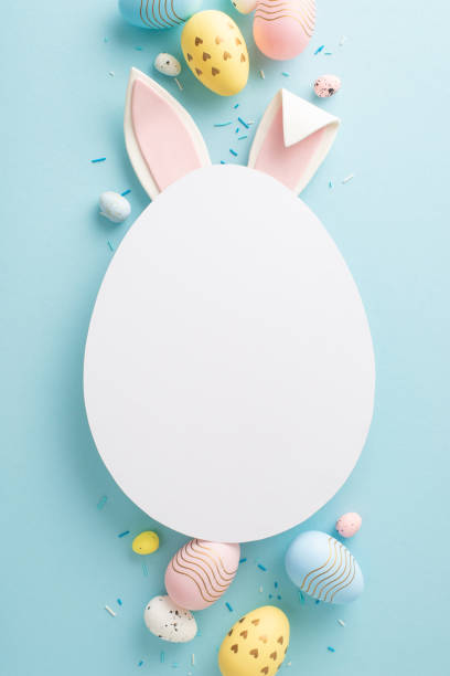 istock Picture-perfect Easter setup: vertical top view of vibrant eggs, sprinkles and rabbit ears emerge from an egg-shaped cutout on a pastel blue background, inviting personalized greetings or ads 2045457179