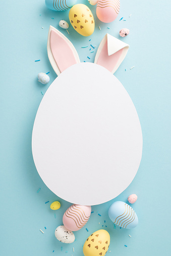 Picture-perfect Easter setup: vertical top view of vibrant eggs, sprinkles and rabbit ears emerge from an egg-shaped cutout on a pastel blue background, inviting personalized greetings or ads
