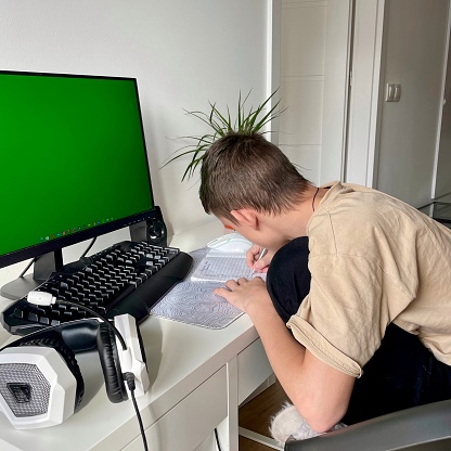 A teenage boy does homework in front of a computer monitor.