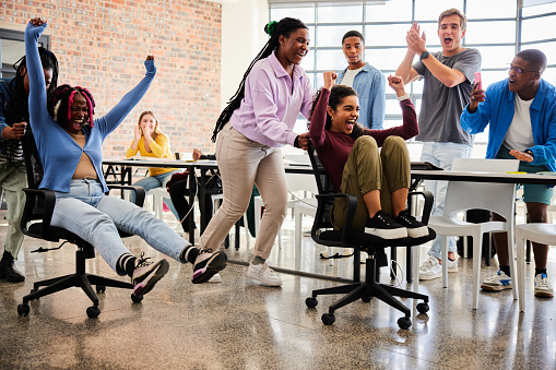 Laughing group of diverse young college students having a chair race around desks in a classroom during a break between lessons