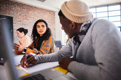 Two diverse young college students talking while sitting together at a table during a class at school with other students