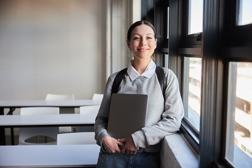 Portrait of a confident young female college student smiling while standing with a laptop by a classroom window