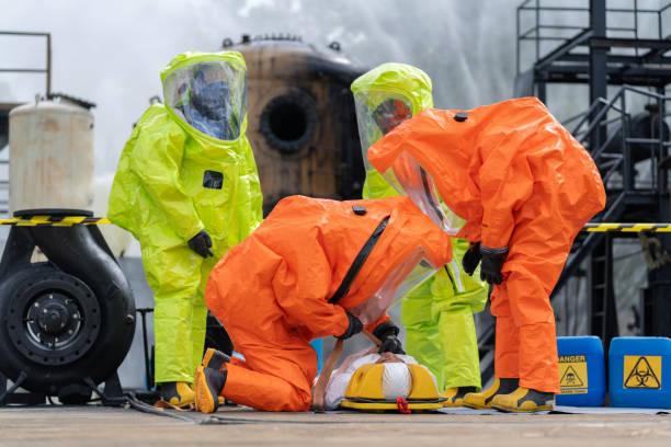team of worker, clad in protective gear including a gas mask and hazmat suit, rushes a patient on a field bed away from the hazardous area surrounding a chemical spill from a factory. teamwork involved in the evacuation are palpable. - radiation protection suit toxic waste protective suit cleaning photos et images de collection