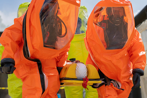 Team of evacuation workers, equipped in safety gear and chemical protection equipment, is captured taking patients out of a chemical plant amidst an urgent chemical spill. Collaborative effort involved in safeguarding lives during a hazardous incident. Exemplify a commitment to safety and protection against the potential dangers of toxic chemicals.