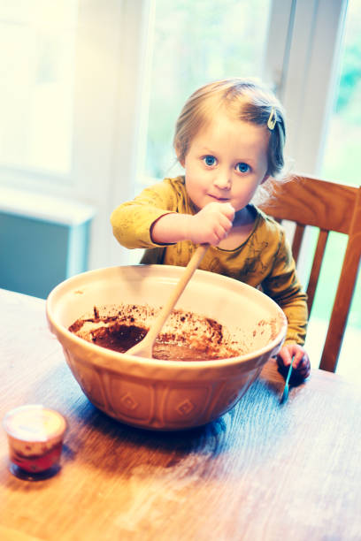 istock Cute 3 year old girl, making her first chocolate cupcakes at a kitchen table mixing the ingredients with a wooden spoon in a ceramic bowl 2045629162
