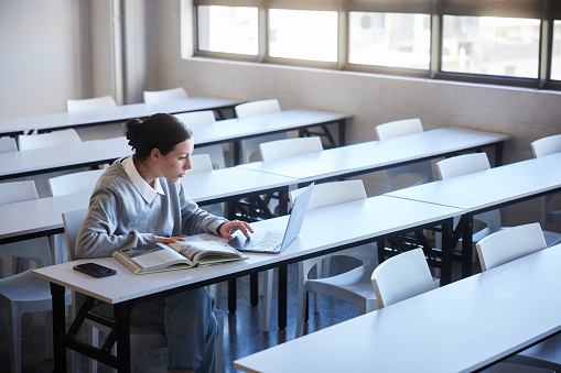 Focused young female college student working on a laptop and reading a textbook while doing her homework at a desk in a classroom